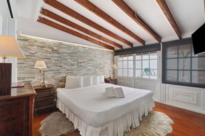 A bedroom with a large bed, wooden beams on the ceiling, a stone wall, and a window with blinds. - Gran Penthouse Luxury The 307 Parque Royal 1 Wiffi y Jacuzzi (Adeje)