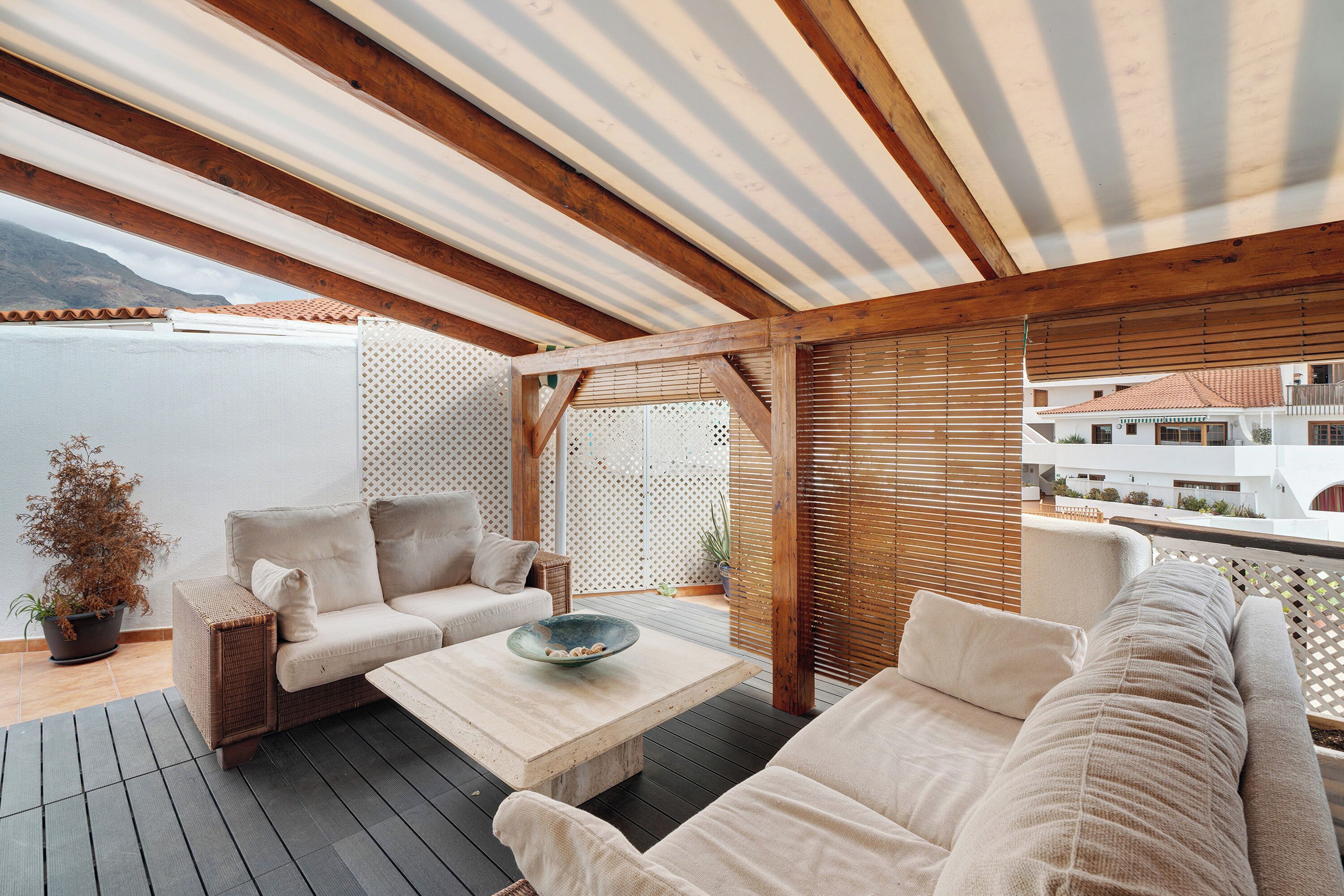 Une terrasse extérieure dotée d'un plafond en bois, de meubles en osier et offrant une vue sur les bâtiments en arrière-plan.