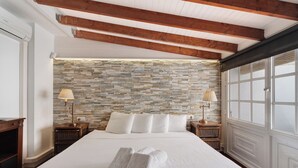 A neatly made bed with white linens and towels, a stone-patterned wall, wooden beams, and a large window with blinds. - Gran Penthouse Luxury The 307 Parque Royal 1 Wiffi y Jacuzzi (Adeje)