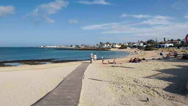 Ein Strand mit einem hölzernen Steg, der zum Wasser führt, Menschen, die sonnenbaden, und Häuser im Hintergrund.