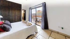 Un dormitorio con cama, armario de madera, baño visible a través de una puerta abierta y vista a una mesa de ping pong exterior por una puerta de vidrio.