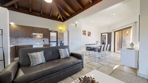 Un espacio habitable moderno con un sofá de cuero negro, una zona de comedor con una mesa y sillas blancas, y una cocina con armarios de madera y salpicadero de azulejos.