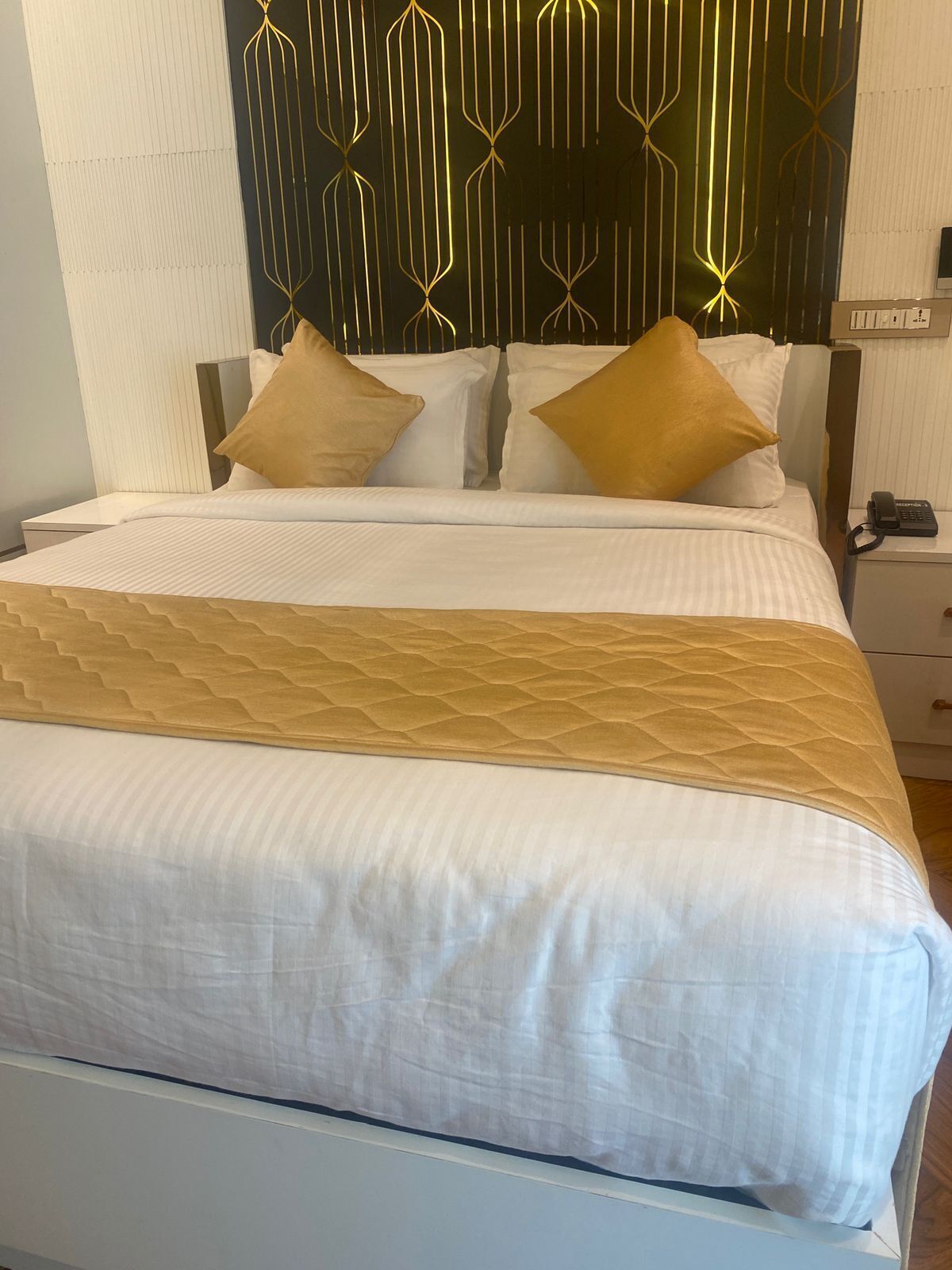 A neatly made bed with a patterned bedspread and two pillows, a bedside table with a phone, and a wall with a geometric light fixture.