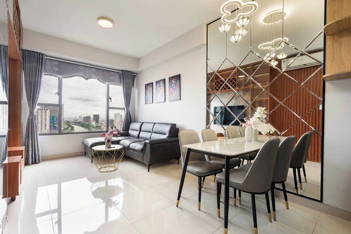 Una sala de estar moderna con sofá, mesa de comedor y sillas. Se aprecia un ventanal con vistas a un paisaje urbano.