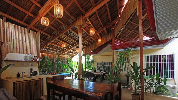 A spacious indoor area with a high ceiling, wooden beams, and hanging light fixtures. There is a long wooden table with chairs, surrounded by potted plants. - Yellow Banana Siargao (General Luna)
