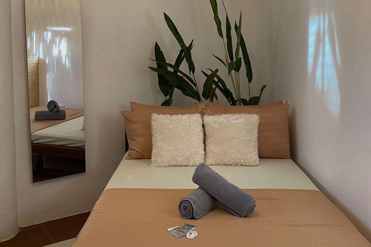Ein kleines, ordentlich eingerichtetes Schlafzimmer mit Bett, Kissen, Spiegel und einer Pflanze.