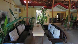 A spacious lobby with a high ceiling, wooden beams, and a central table. There are two dark-colored sofas with patterned cushions and several potted plants. - Yellow Banana Siargao (General Luna)