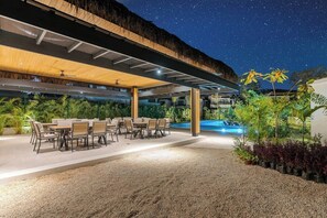 An outdoor dining area with tables and chairs, a pool, and a thatched roof structure.
