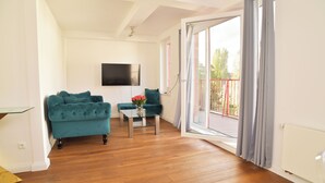 Una sala de estar con un sofá verde azulado, una mesa de centro de vidrio con flores y un televisor en la pared.