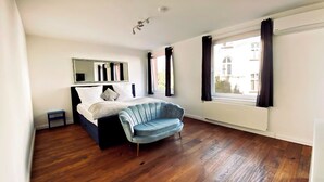 Un dormitorio con una cama, un sillón azul, una mesita, un espejo y una ventana con cortinas.