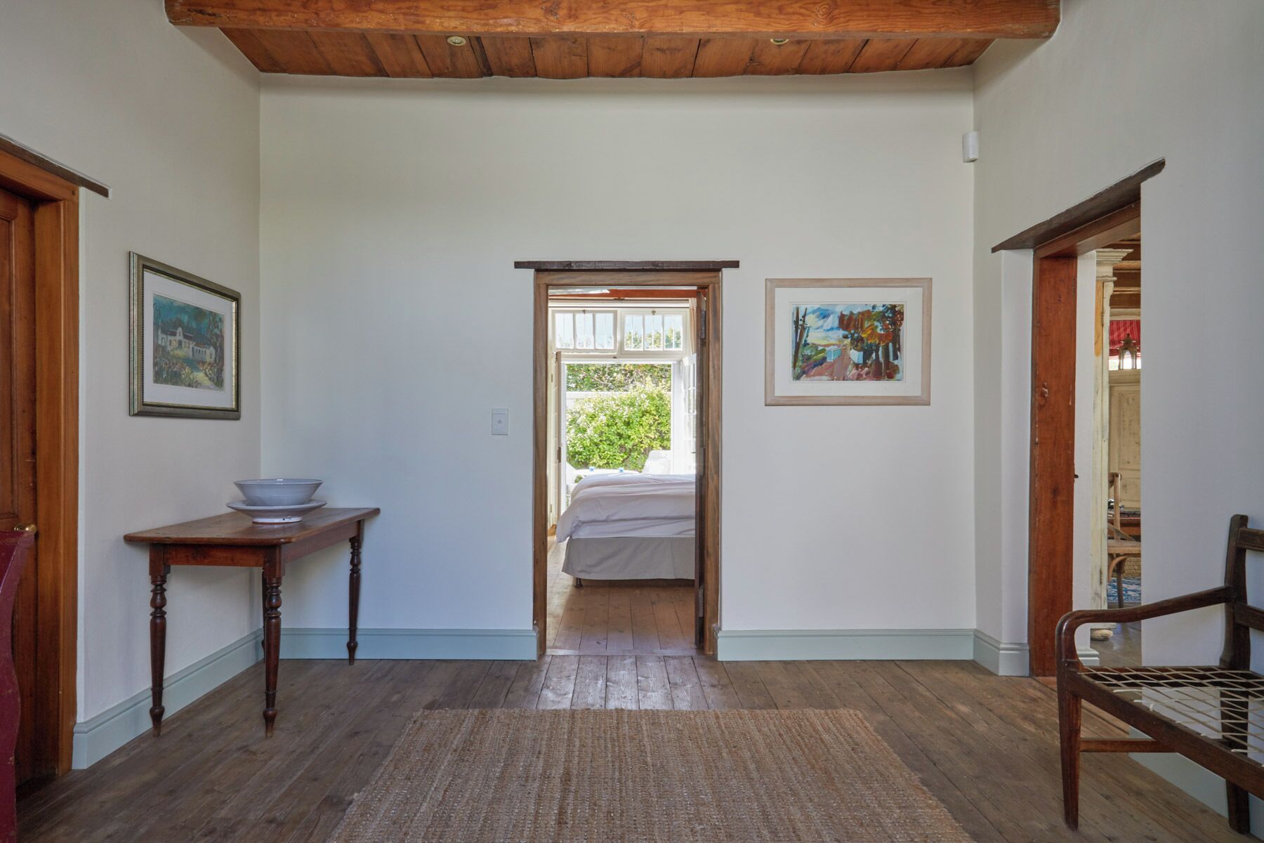 A room with a wooden floor, a bed, a table with a bowl, and two framed pictures on the walls.