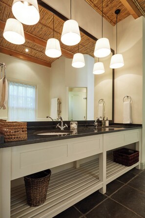 Un baño moderno con encimera negra, muebles de color blanco y techo de madera. Hay dos lavamanos, un espejo y lámparas colgantes.