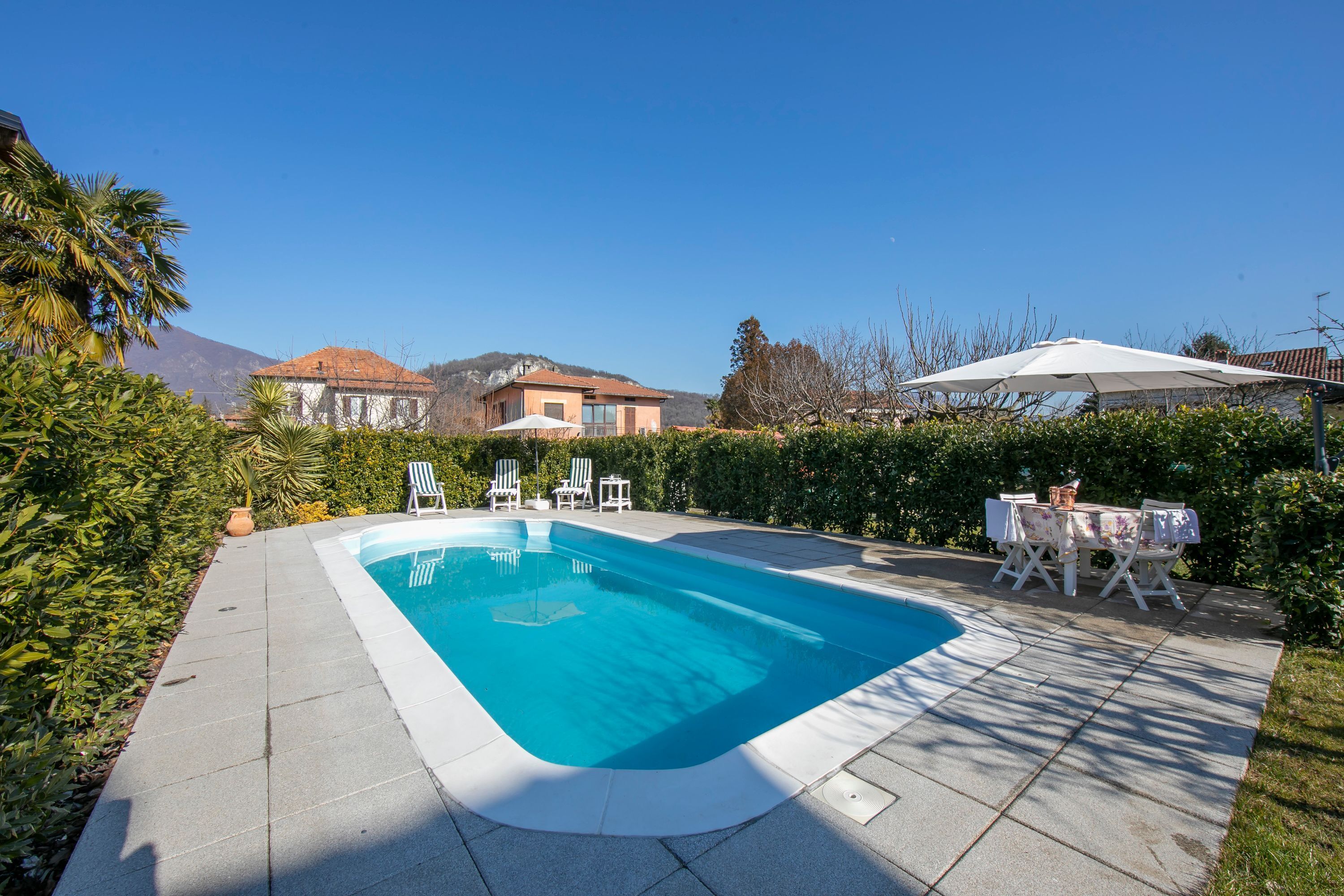 Ein rechteckiger Swimmingpool mit weißem Rand, umgeben von einer gepflasterten Terrasse. Weiße Liegestühle und ein Tisch mit einem weißen Sonnenschirm stehen bereit. Im Hintergrund sieht man ein Haus mit rotem Dach und einen Berg.