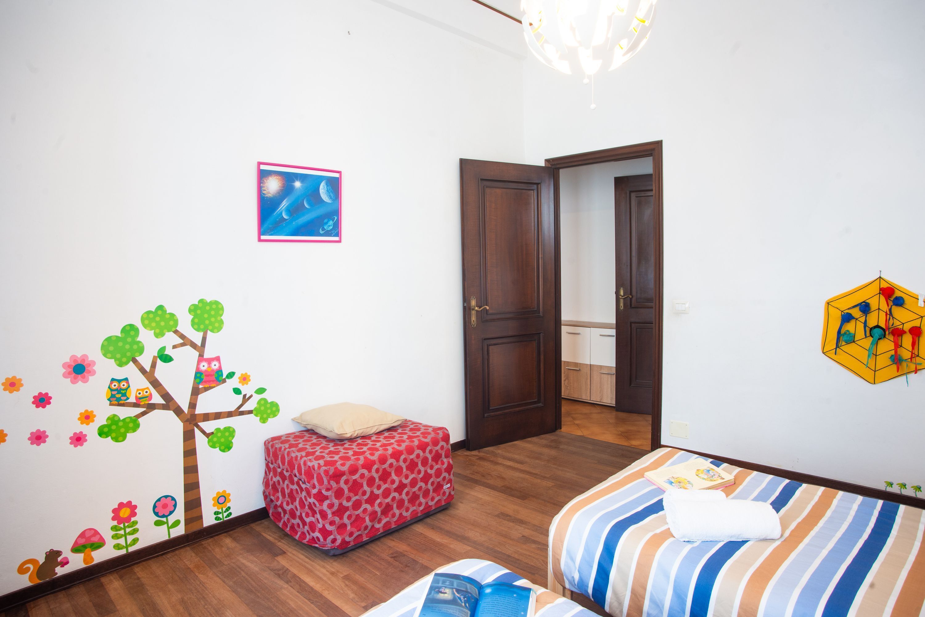 A child's room with a bed, a red ottoman, a wall decal of a tree with flowers and owls, and a door leading to another room.
