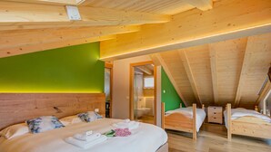 Un dormitorio con una cama grande, dos camas más pequeñas, vigas de madera, una pared verde y un baño visible a través de una puerta abierta.