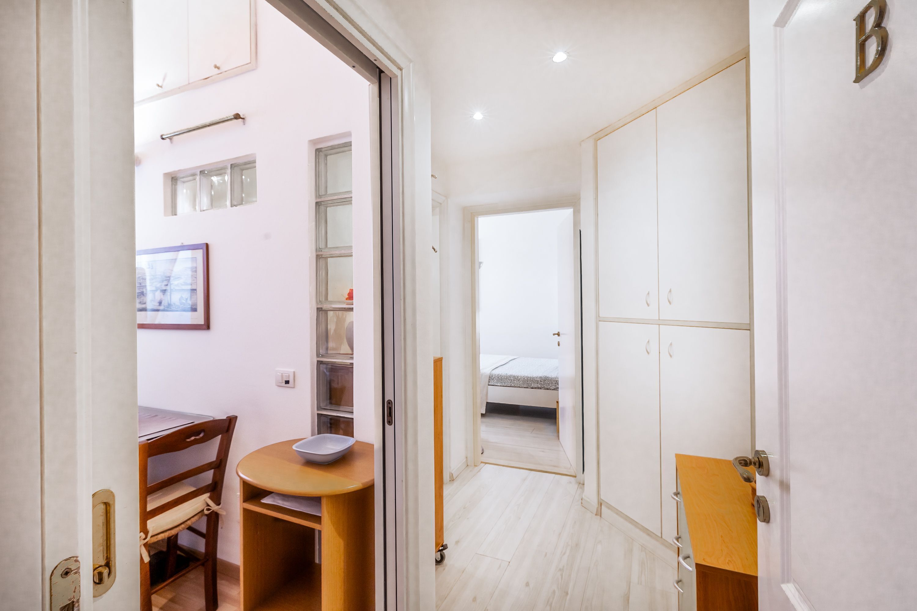 A narrow hallway with a wooden table and chair, a small round table with a bowl, and a bed in the adjacent room.