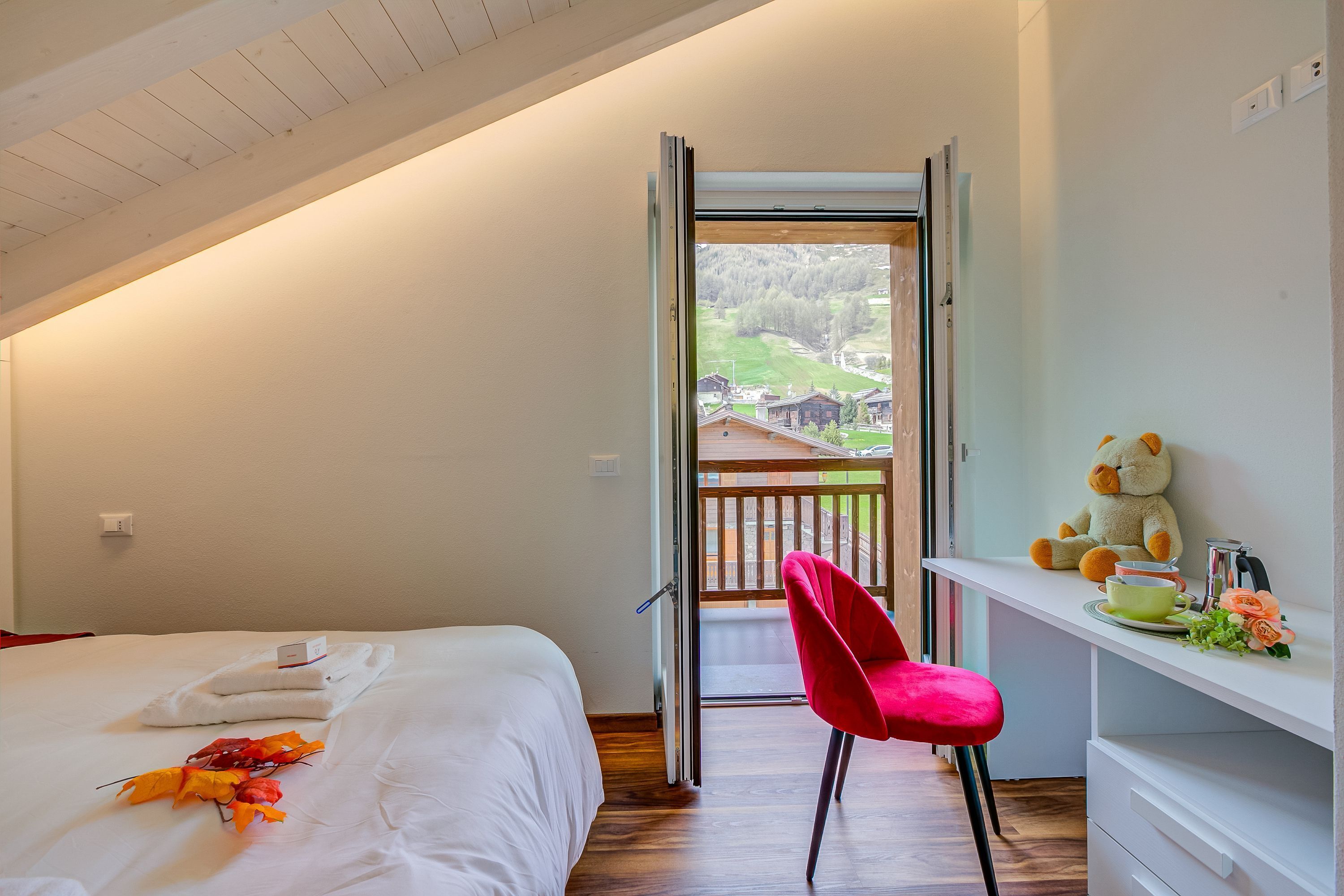 Et soveværelse med en seng, et skrivebord med en bamse og udsigt til en balkon.