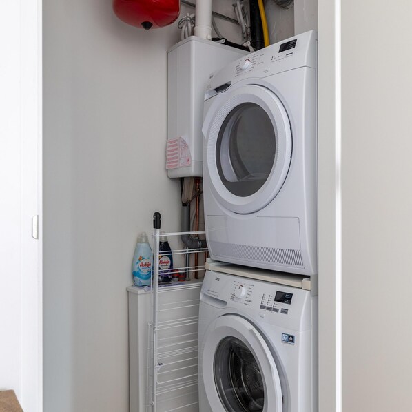 Una lavadora y secadora apiladas con un panel de control, una etiqueta de advertencia de seguridad y un estante con productos de limpieza.
