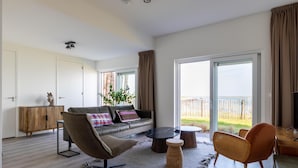 A modern living room with a sofa, coffee table, and chairs, a wooden cabinet, and large windows offering a view of the outdoors. - Lux Apvm Lagunab-havenw 8-32 S-annaland (Sint-Annaland)
