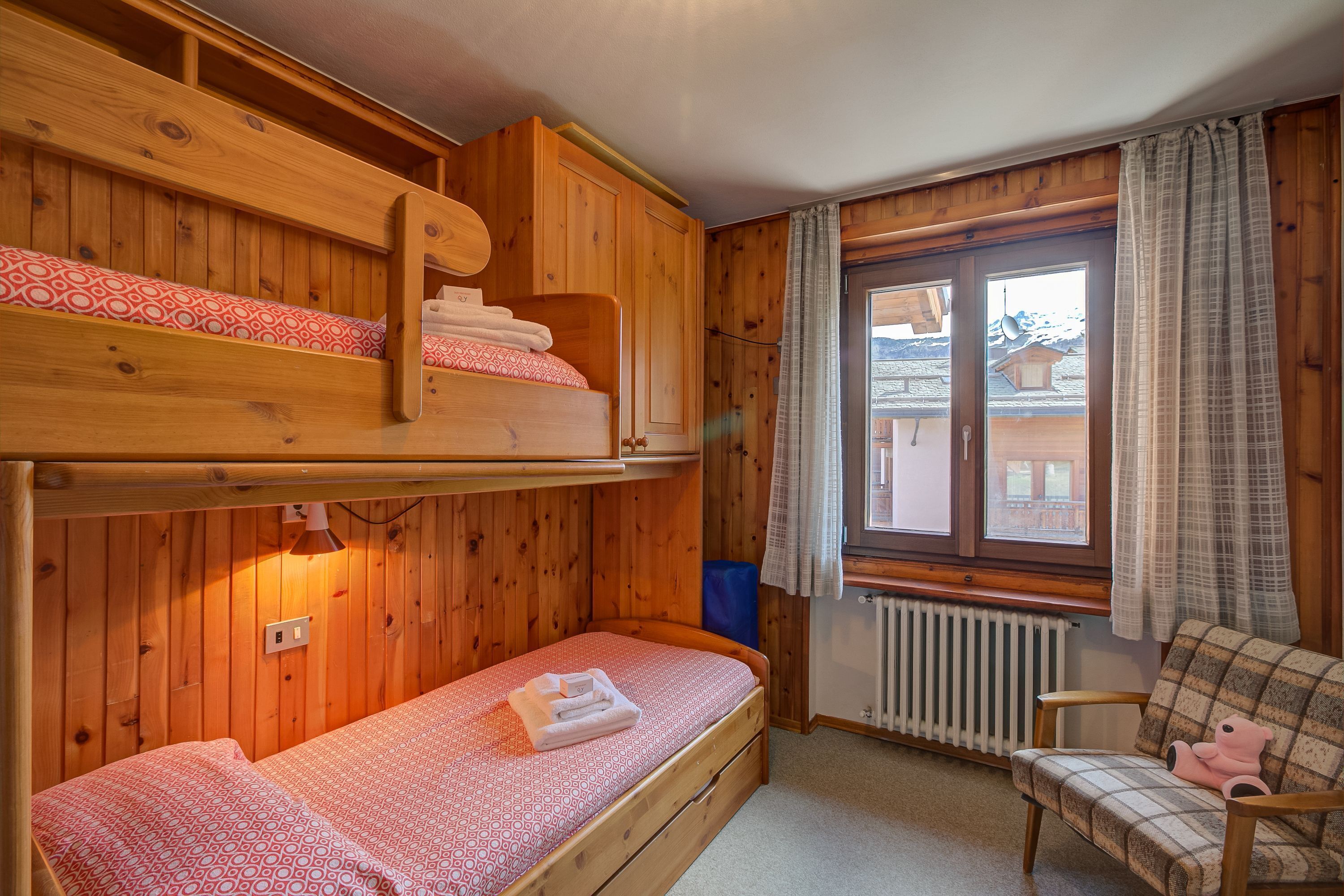A wooden cabin room with bunk beds, a chair, and a window with curtains.