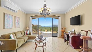 A spacious living room with a sofa, a coffee table, a TV, and a balcony with a scenic view. - Majestic Oceanview Condo with King Beds (Playas Del Coco)