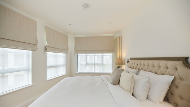 Quarto de hotel com uma cama grande, cortinas bege e uma luminária de cabeceira.