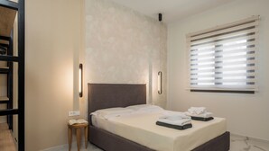 A modern bedroom with a large bed, a bedside table with a book, and a window with blinds. - Orama Luxury Apartments (Chania)