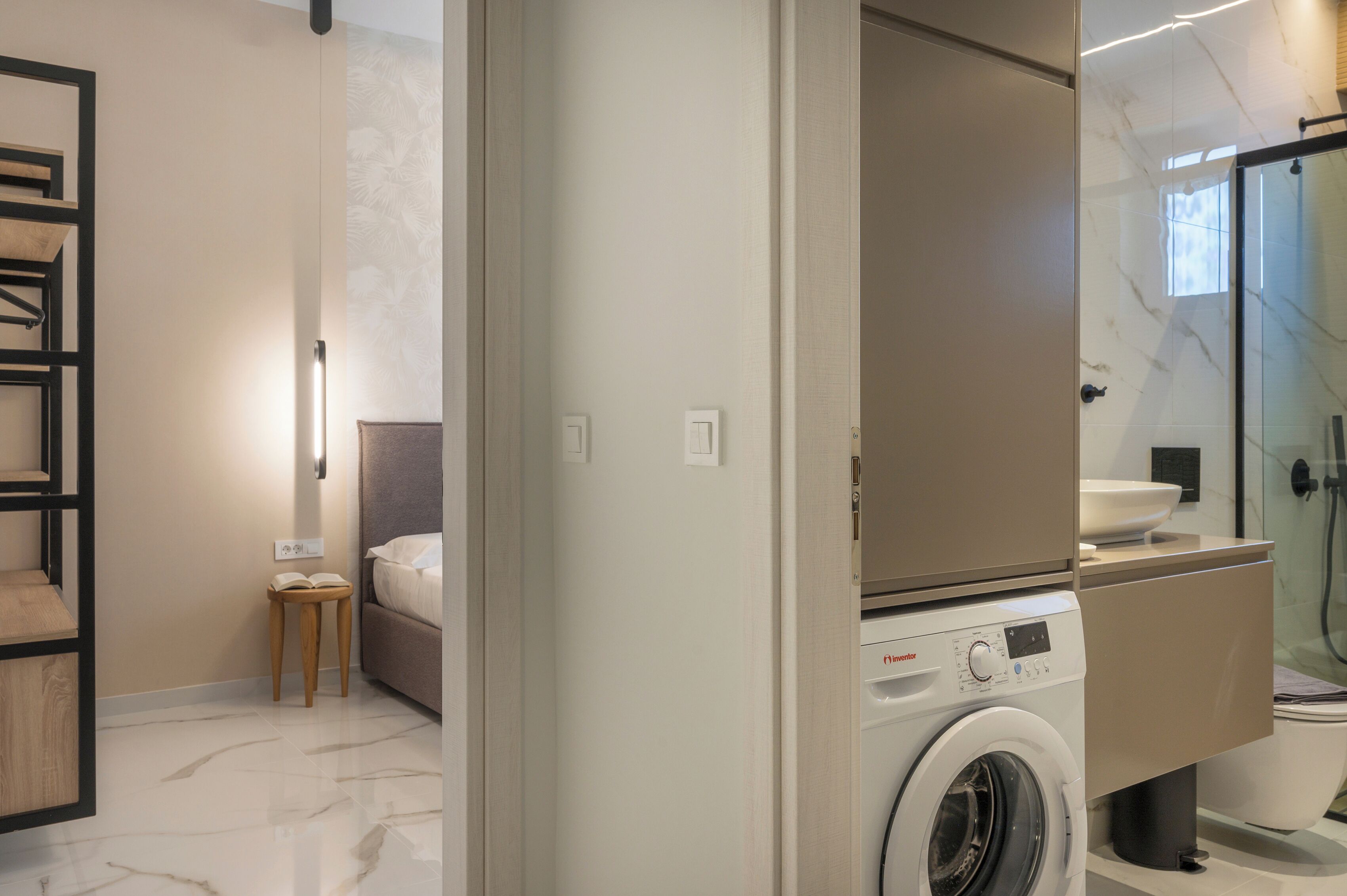 A modern bathroom with a built-in washing machine, a glass shower enclosure, and a bedside table with a book.