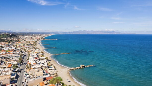 An aerial view of a coastal city with a long beach, clear blue sky, and distant mountains. - Orama Luxury Apartments (Chania)