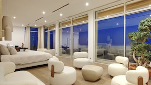 A modern bedroom with a large bed, multiple white armchairs, a potted plant, and large windows offering a view of the city. - Villa Tigliola (Zonza)