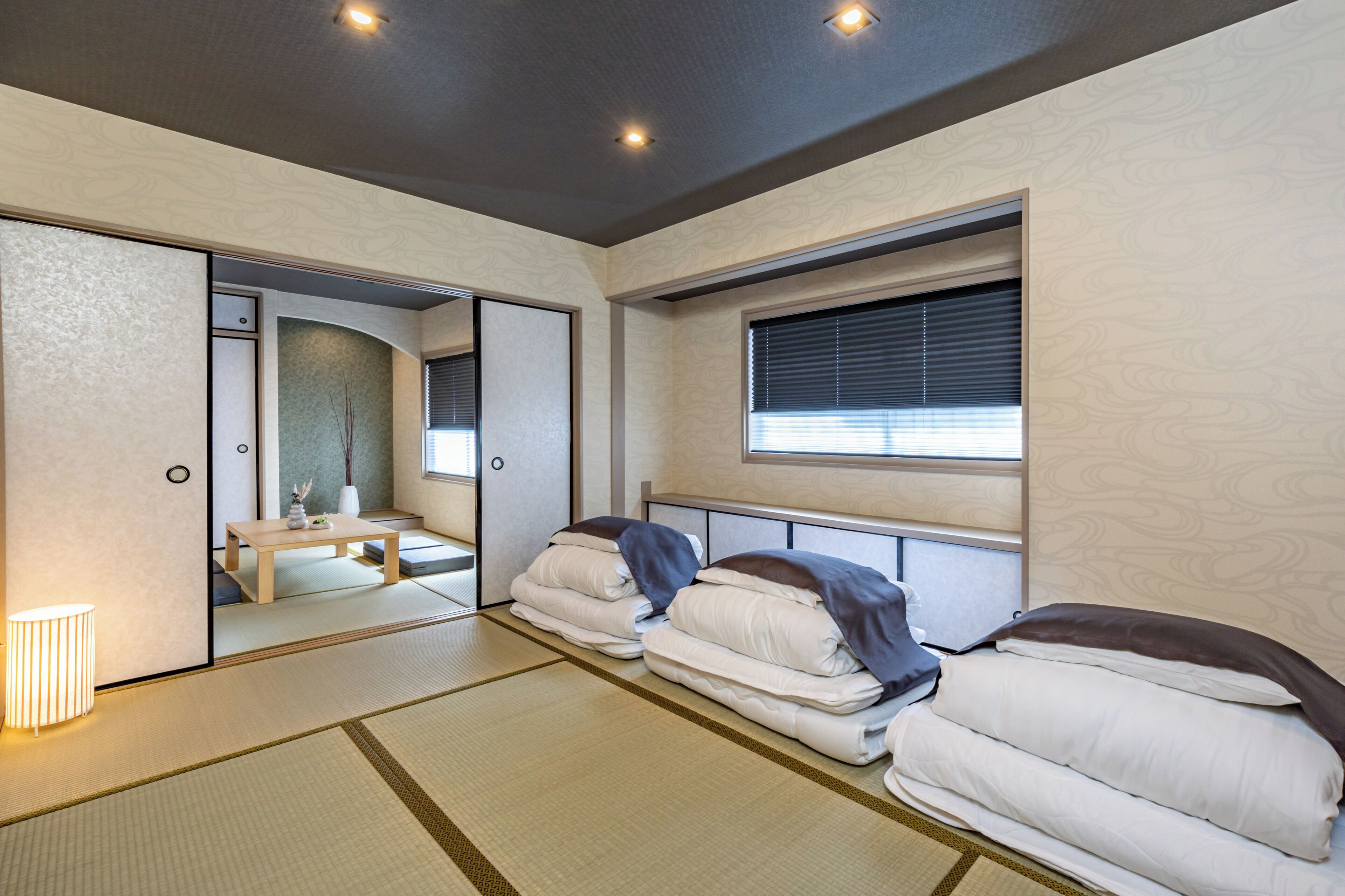 A traditional Japanese-style room with tatami flooring, two beds with white bedding, a small table, and a window with blinds.