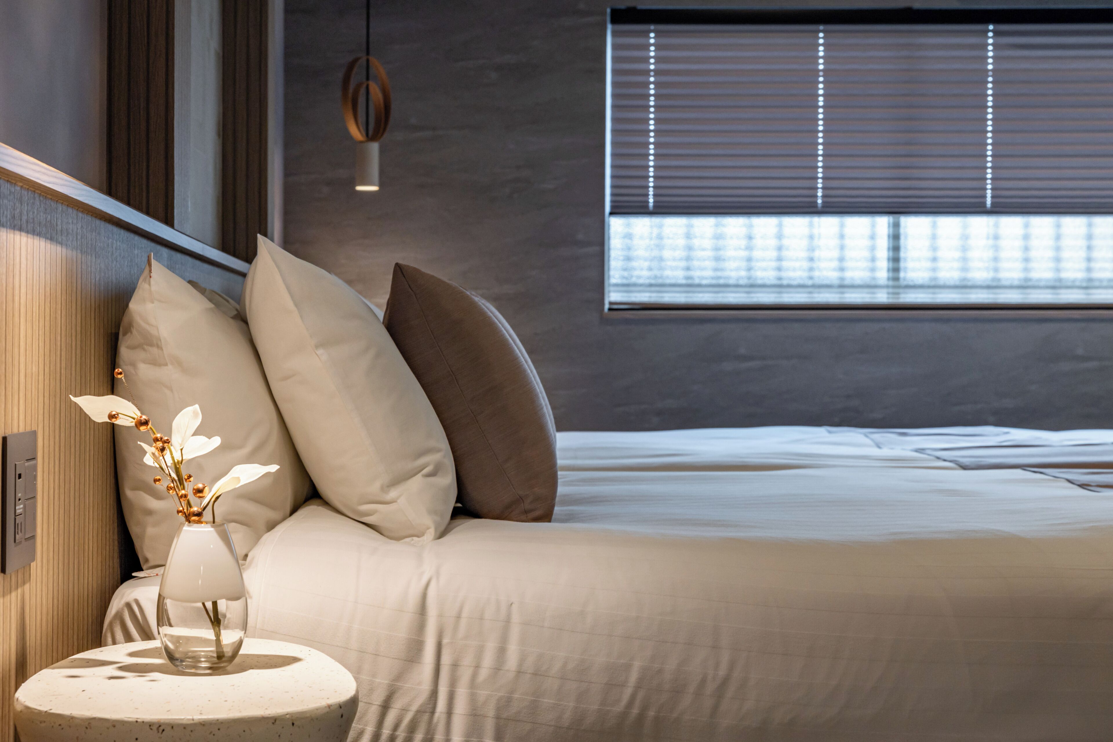 Un dormitorio moderno con cama, almohadas, un jarrón con flores y una mesa redonda.