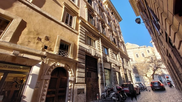 Un edificio histórico con una barbería en la planta baja, scooters y coches estacionados en una calle adoquinada.