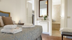 Un dormitorio con cama, mesita de noche, tocador con lámpara, espejo y baño visible a través de una puerta abierta.