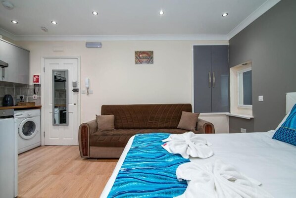A bedroom with a bed, a washing machine, a sofa, and a small kitchen area. - Camden Apartments (London)