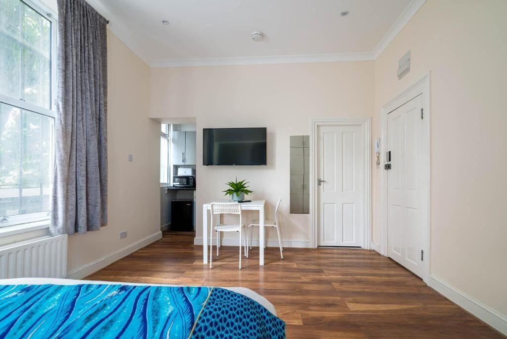 A room with a bed, a TV mounted on the wall, a small table with chairs, and a window with curtains.