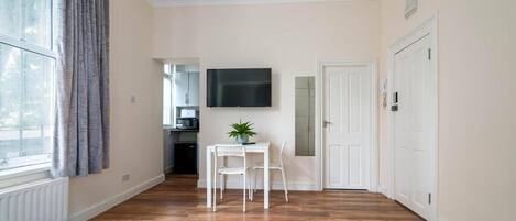 A room with a bed, a TV mounted on the wall, a small table with chairs, and a window with curtains.