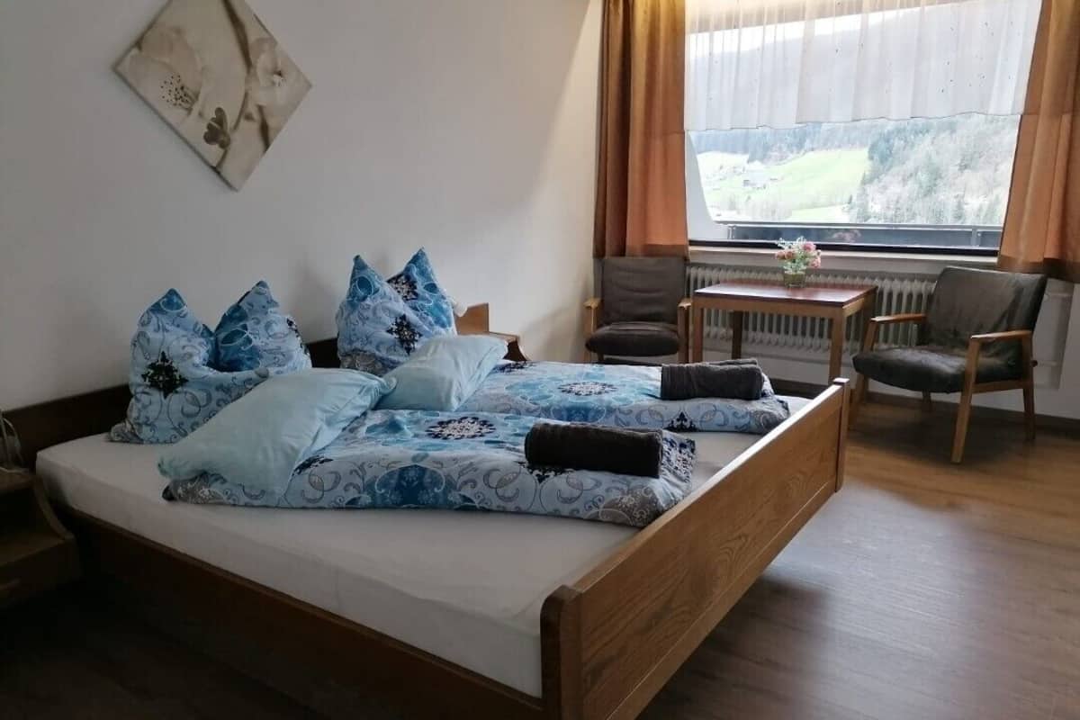 A bedroom with a wooden bed, blue patterned bedding, a chair, a small table, and a window with curtains.