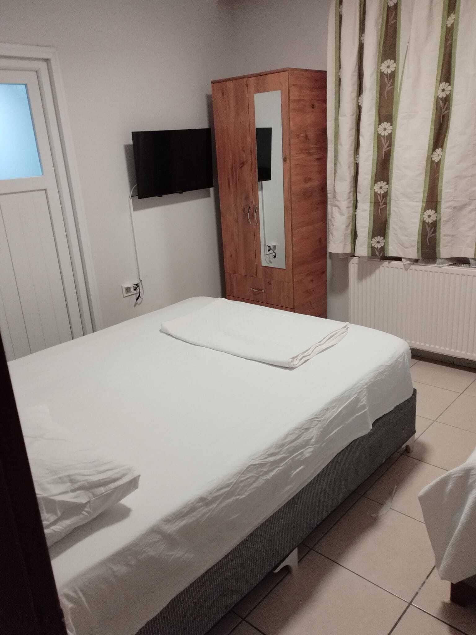 A bedroom with a bed, a wooden wardrobe, a flat-screen TV mounted on the wall, and a window with curtains.