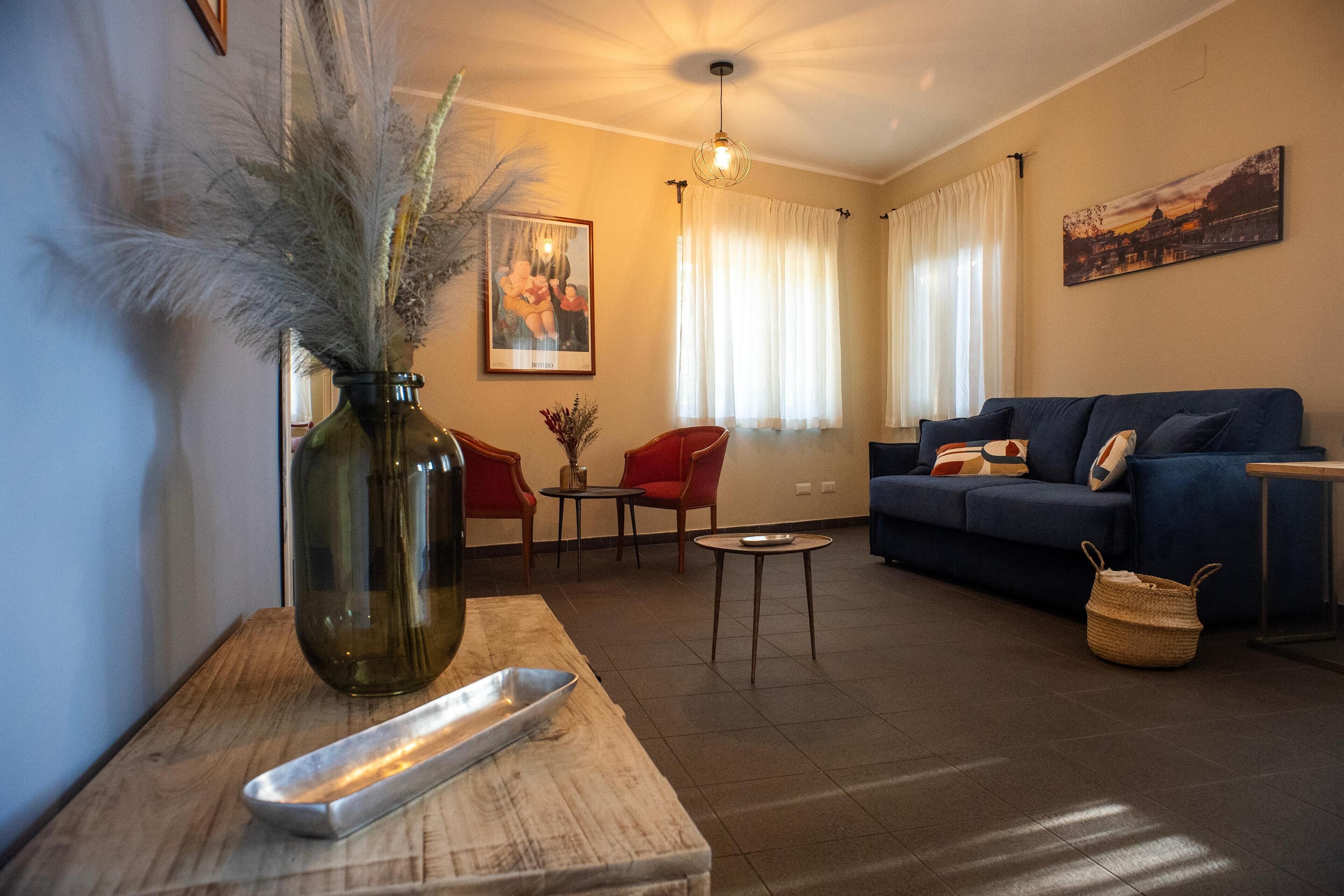 A living room with a sofa, chairs, a coffee table, a vase with dried plants, and a painting on the wall.