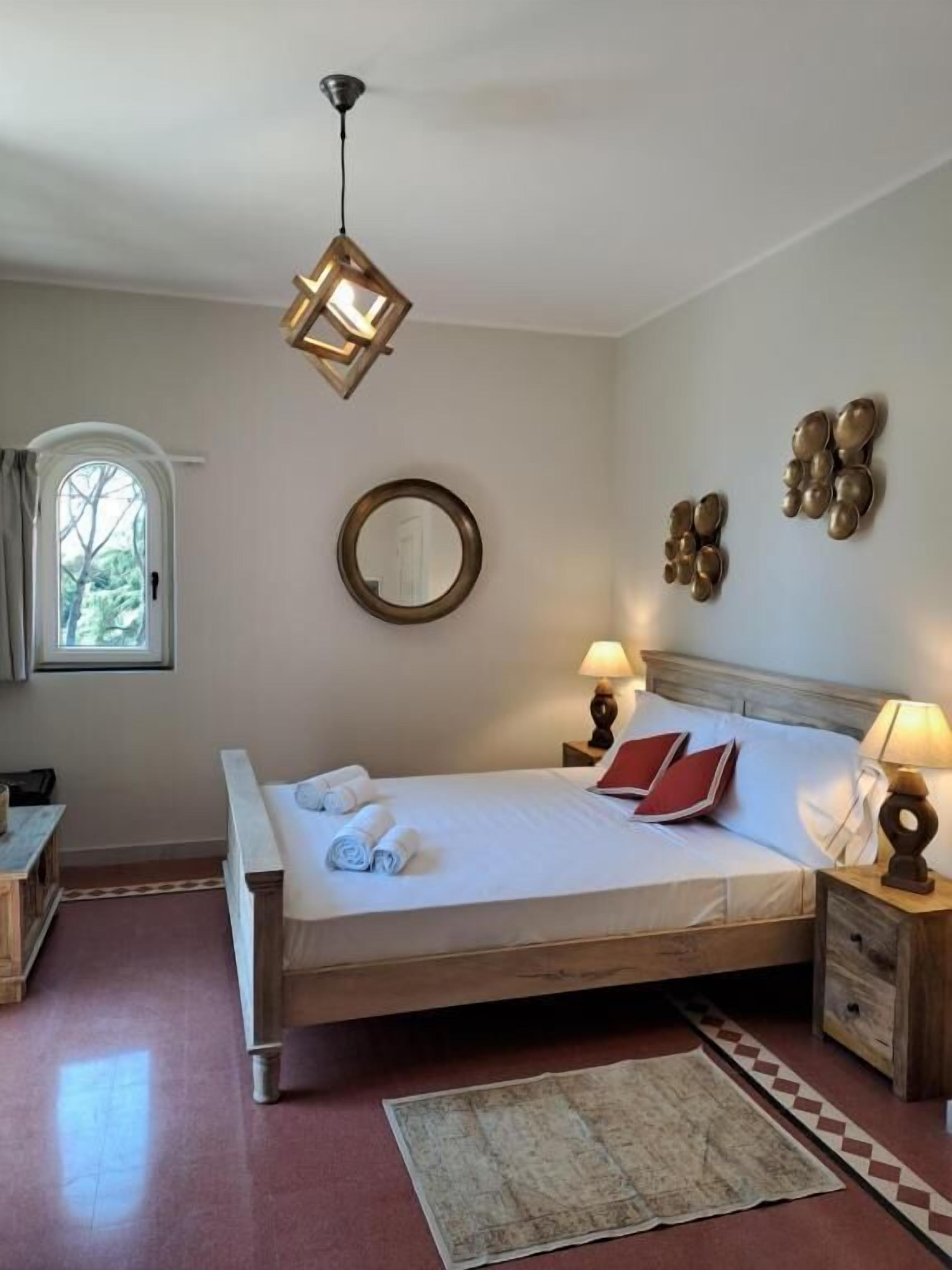 A hotel room with a wooden bed, a round mirror, two wall sconces, and a window with curtains.