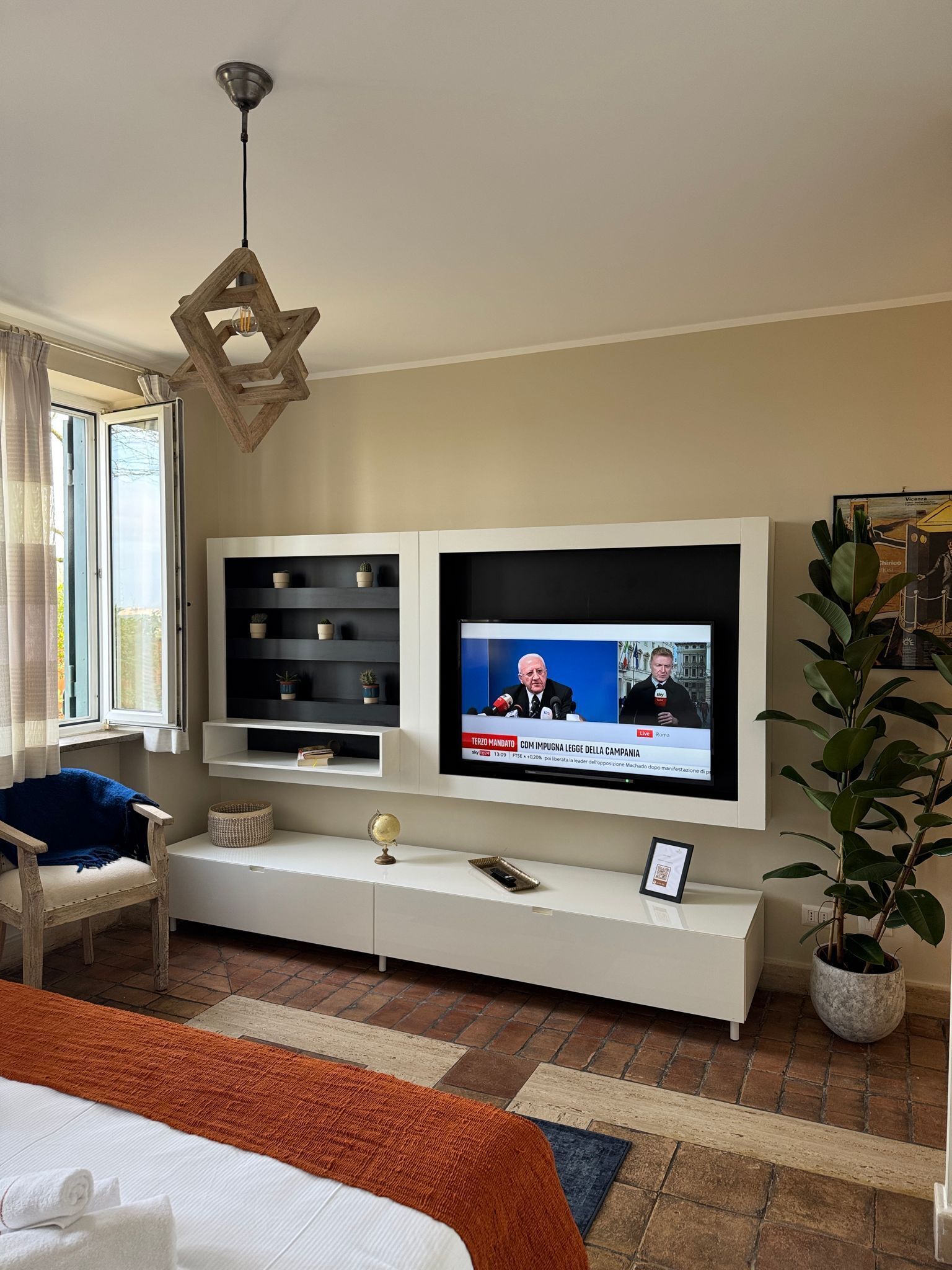 A modern hotel room with a large flat-screen TV mounted on the wall, a wooden chair, a bedside table with a globe and a framed certificate, and a potted plant.