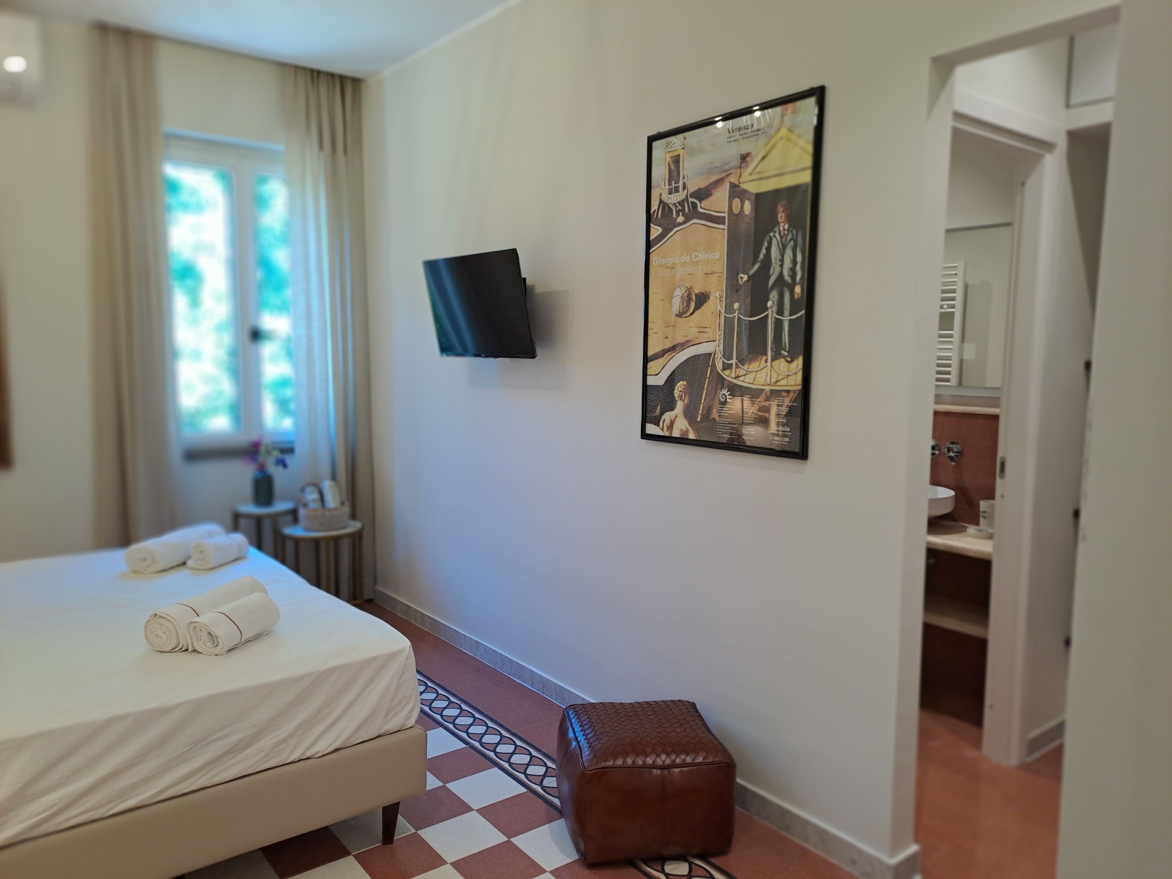 Habitación de hotel con cama, televisor montado en la pared, un cuadro, una mesita con un florero y un baño visible a través de una puerta abierta.