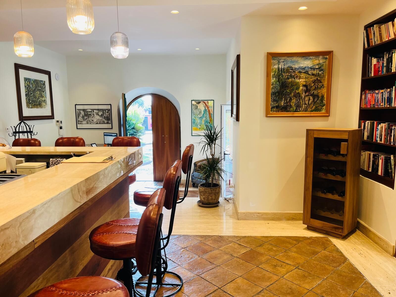 A hotel lobby with a reception desk, bar area, leather chairs, potted plants, artwork, and a bookshelf.