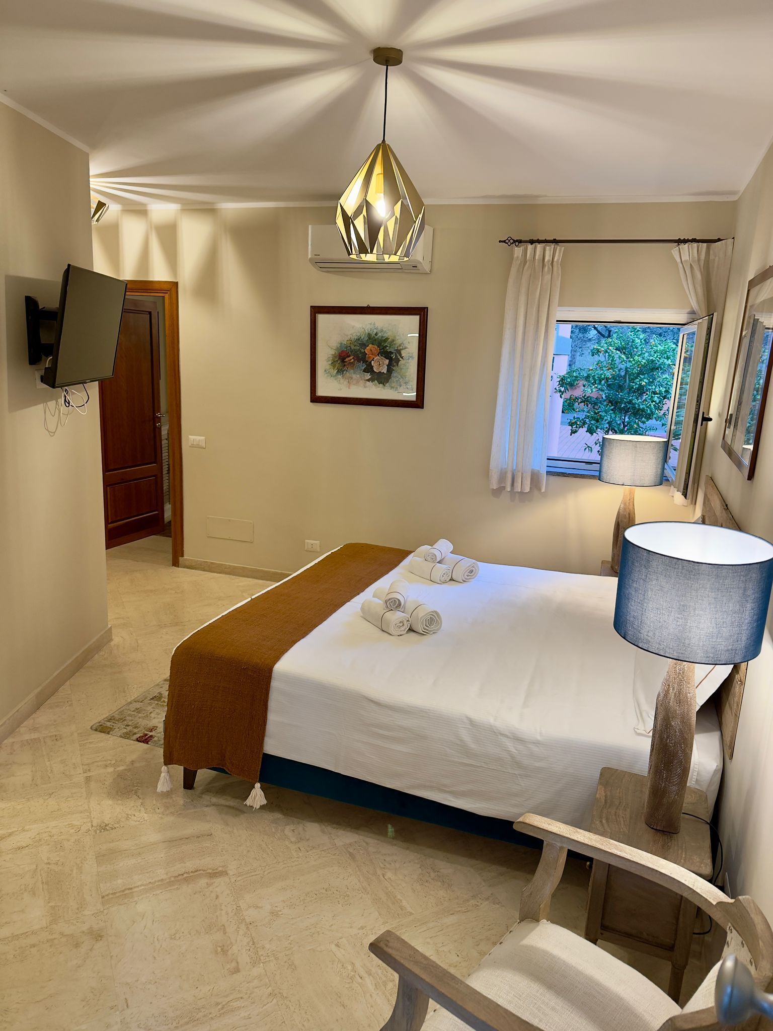 Une chambre d’hôtel avec un lit, un banc en bois, un téléviseur fixé au mur, un luminaire au plafond et une fenêtre avec des rideaux.