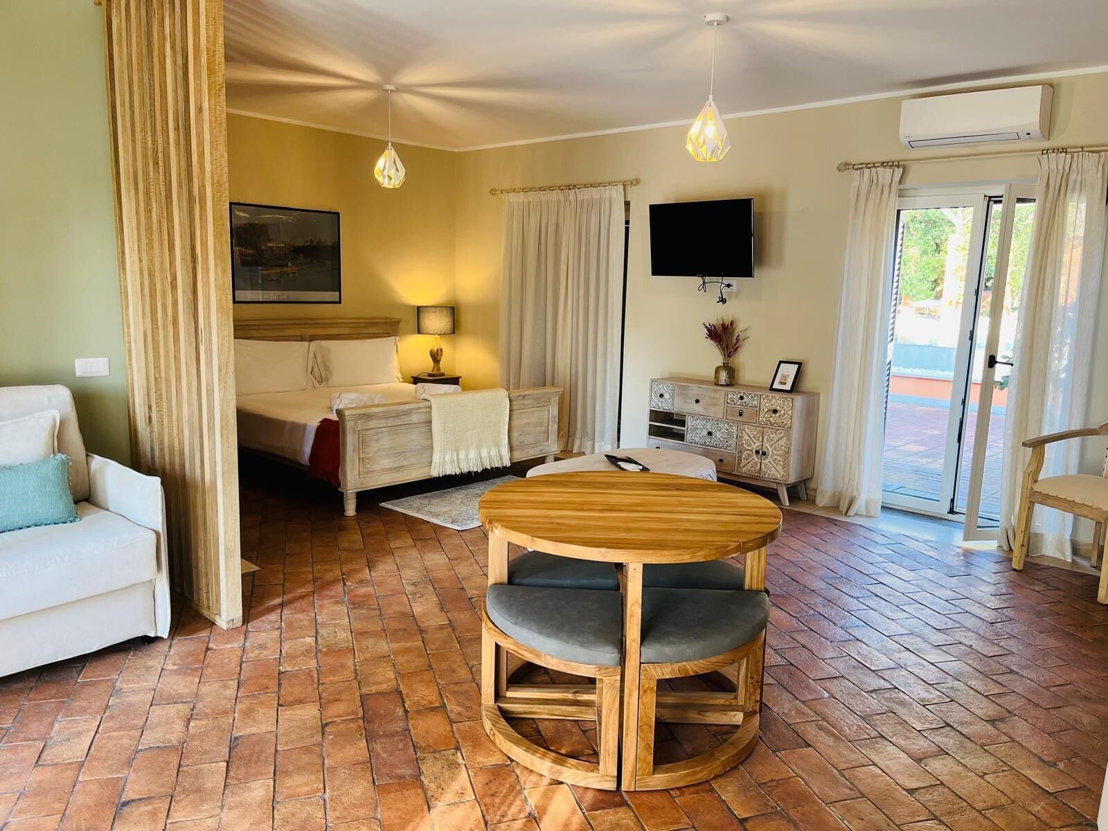 A hotel room with a bed, a TV, a wooden dining table with chairs, a white sofa, and a brick floor.