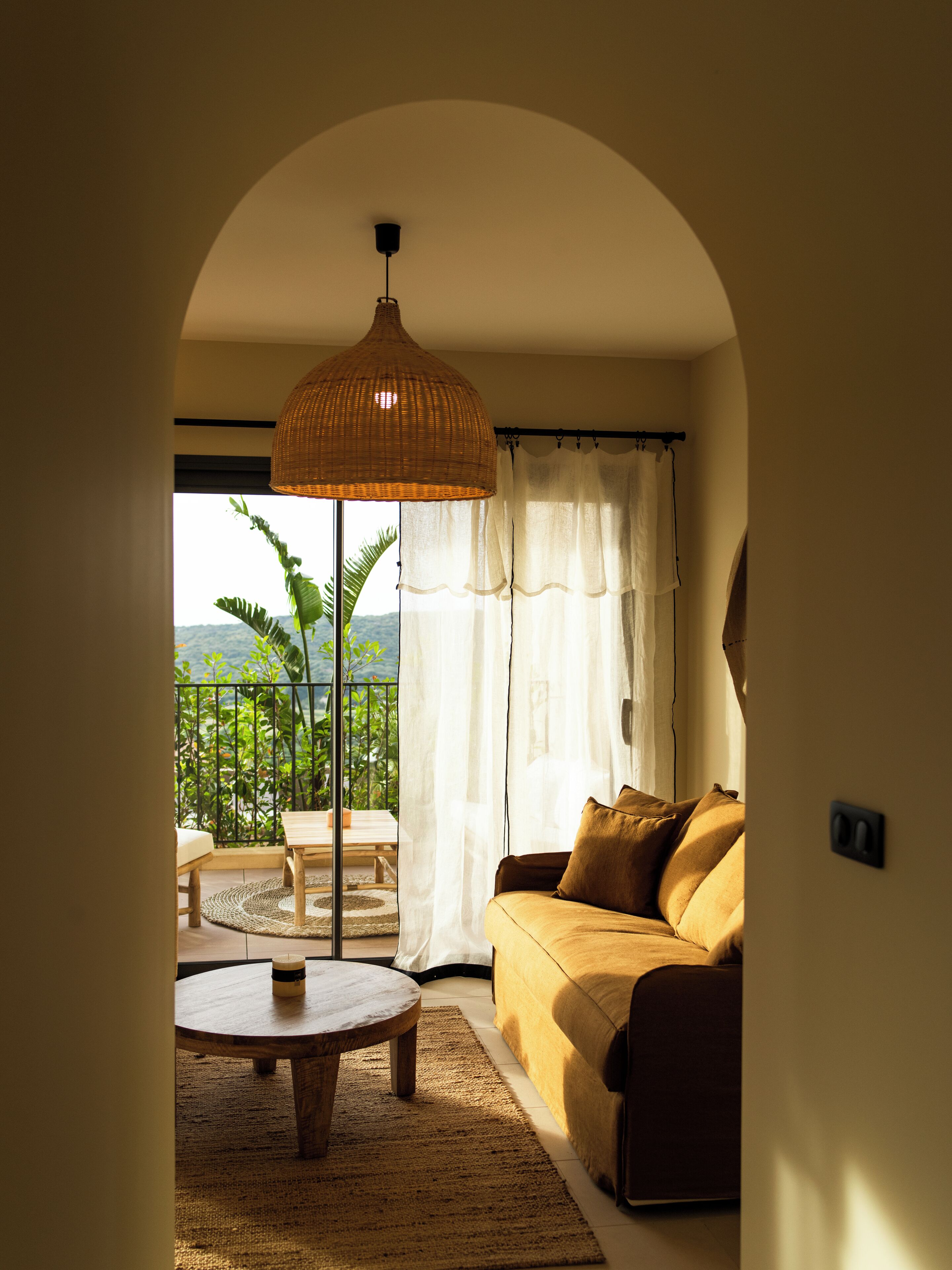 A cozy living room with a sofa, a round coffee table, and a hanging light fixture. A balcony with a view of greenery and a chair is visible through a window.
