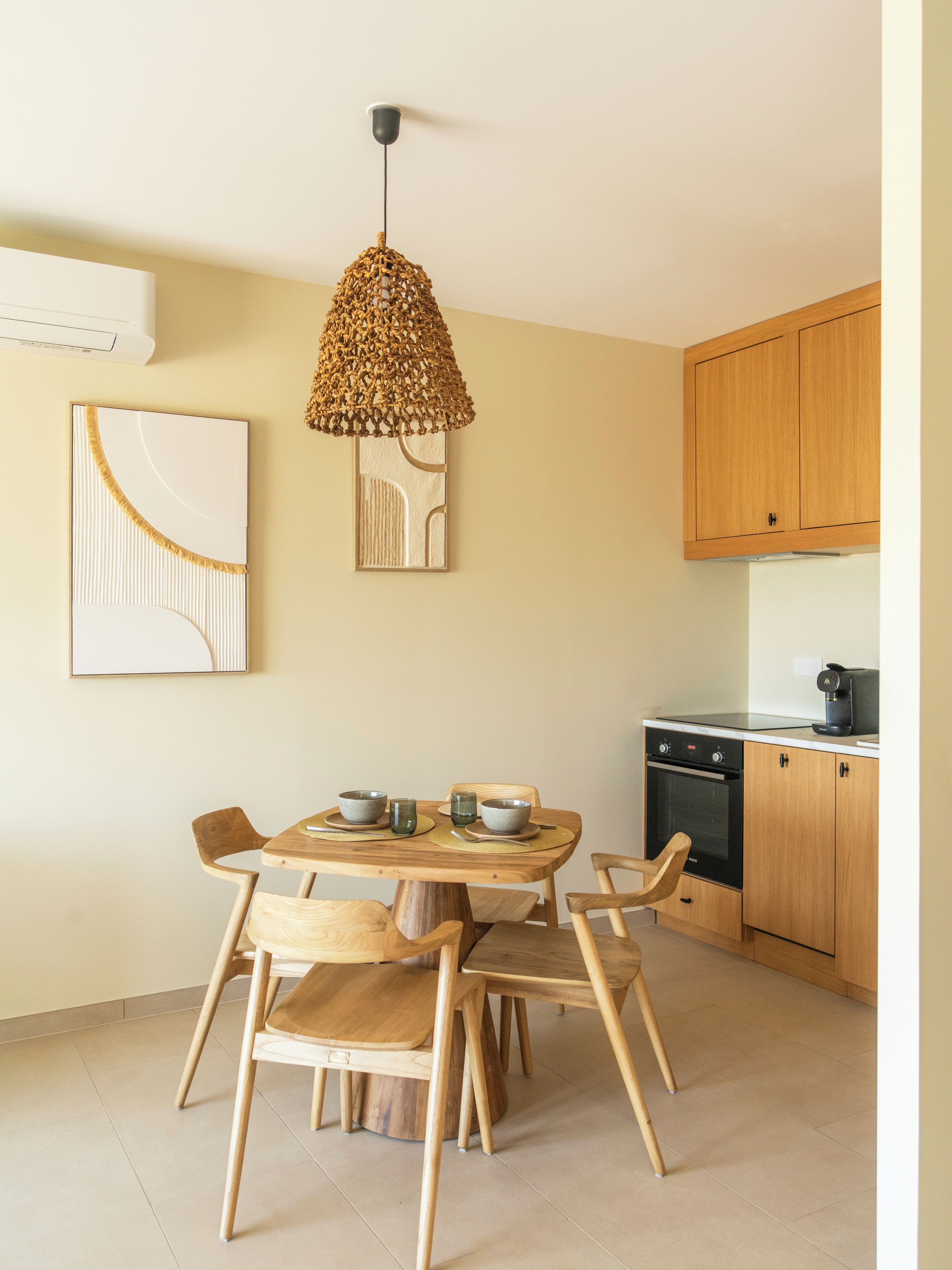 Une cuisine moderne avec une table ronde en bois, quatre chaises, un luminaire suspendu en fibres tressées, une œuvre d’art abstraite accrochée au mur et des armoires en bois.