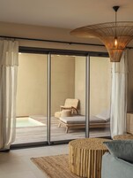 Une chambre moderne dotée d'une porte vitrée donnant sur un balcon, d'un fauteuil en osier, d'un banc rembourré, d'un tabouret en fibres tressées et d'un luminaire suspendu.