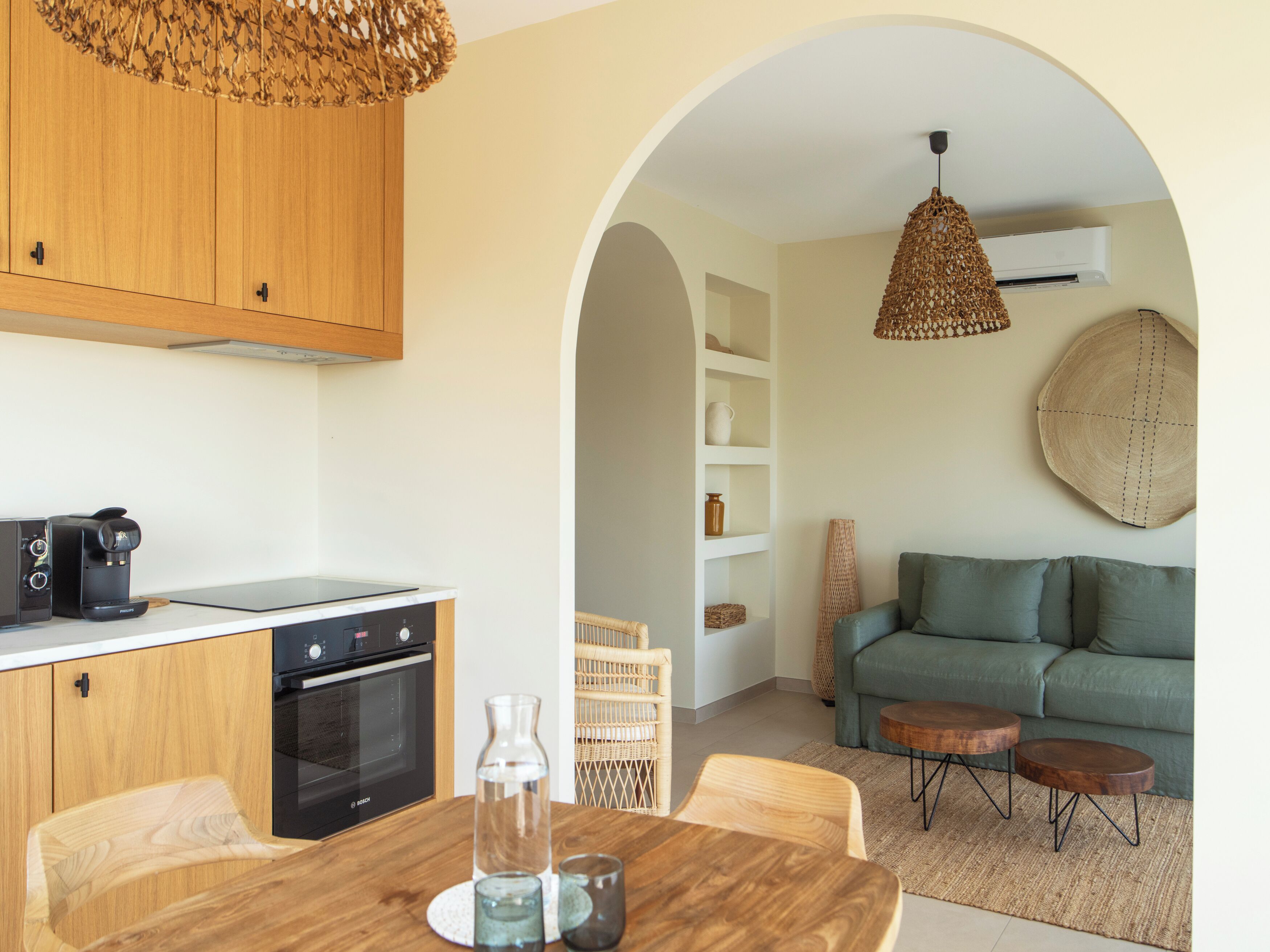 Una cocina moderna con armarios de madera, horno empotrado y cafetera. Hay una mesa de comedor con sillas y un jarrón. La zona de estar cuenta con un sofá verde, una mesa redonda de centro y una lámpara colgante.